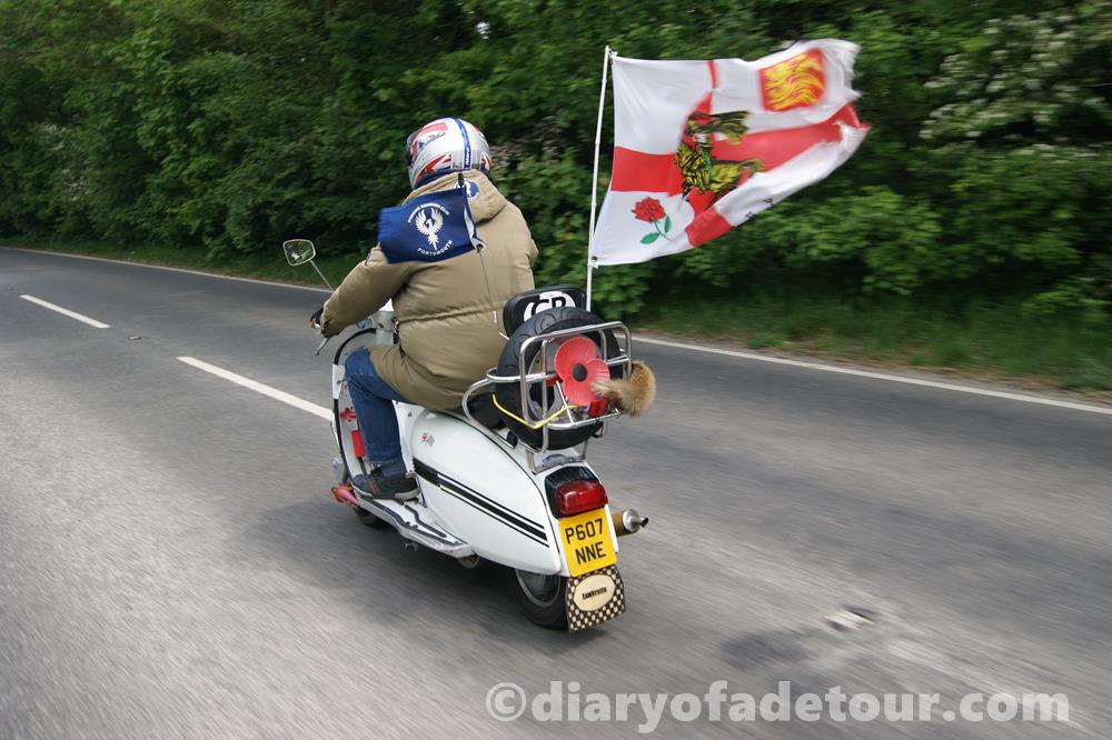 White Lambretta scooter riding along the road with an England flag attached to the back rack