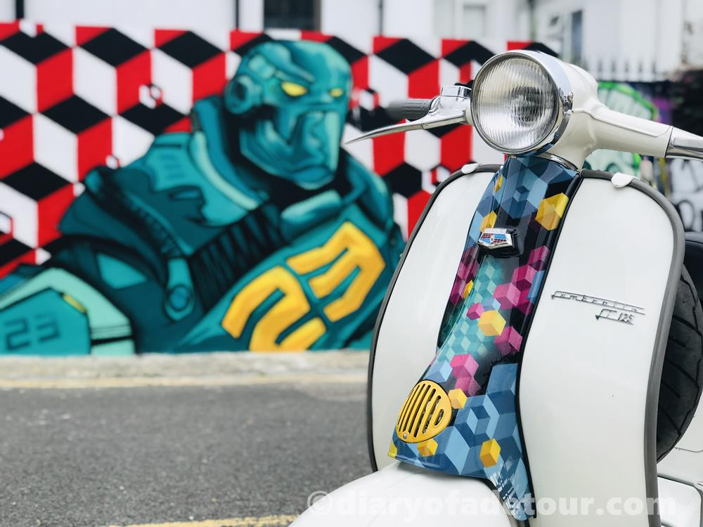 Front view of a classic Lambretta with painted horncover, in front of a mural with a similar design by Snub 23 in Trafalgar Lane, Brighton