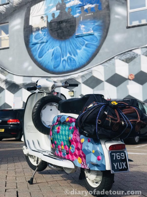 Scooter with street art painted on the side panels in front of a mural in Portsmouth by My Dog Sighs and Snub23