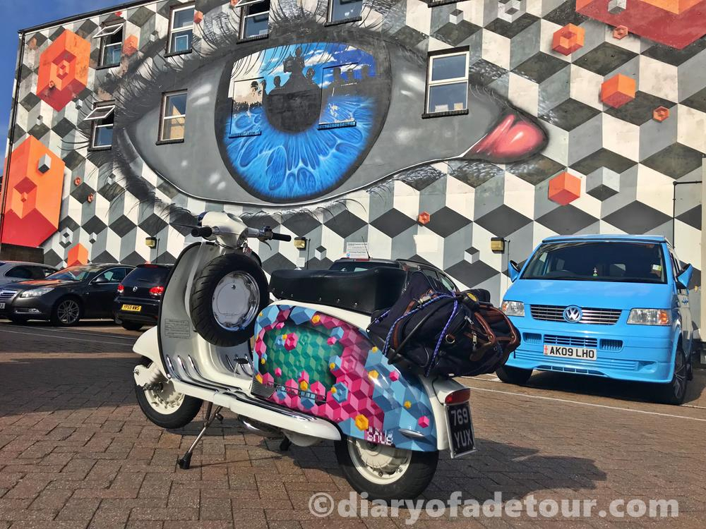 Scooter with street art painted on the side panels in front of a mural in Portsmouth by My Dog Sighs and Snub23