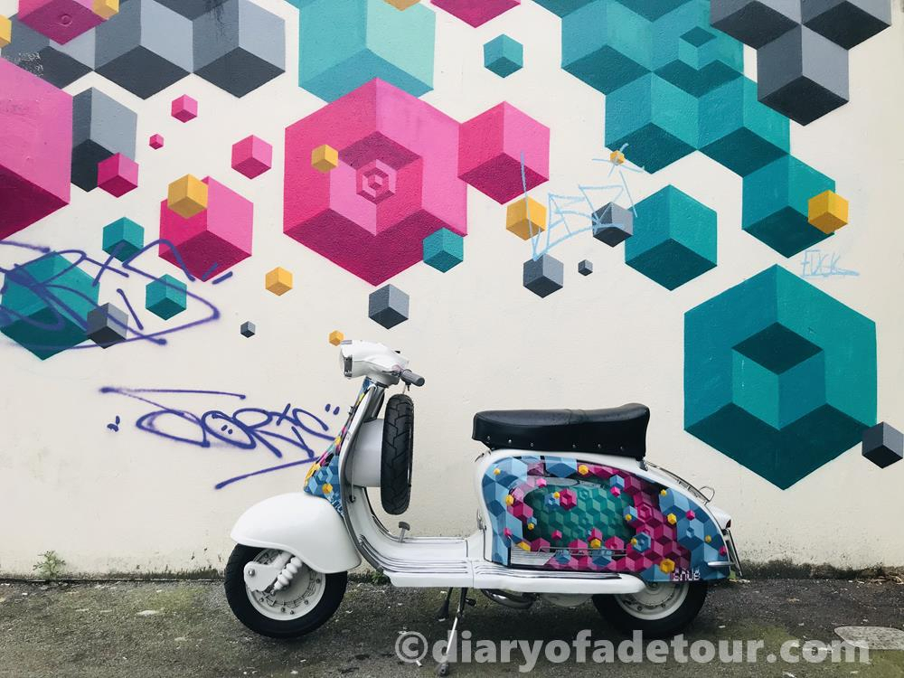 Scooter with colourful isometric painted side panels in front of a wall mural in the same style