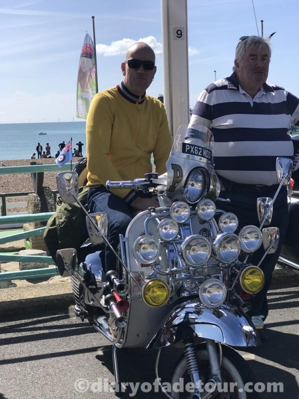 A mod sitting on a classic Vespa with lights and mirrors on Brighton seafront