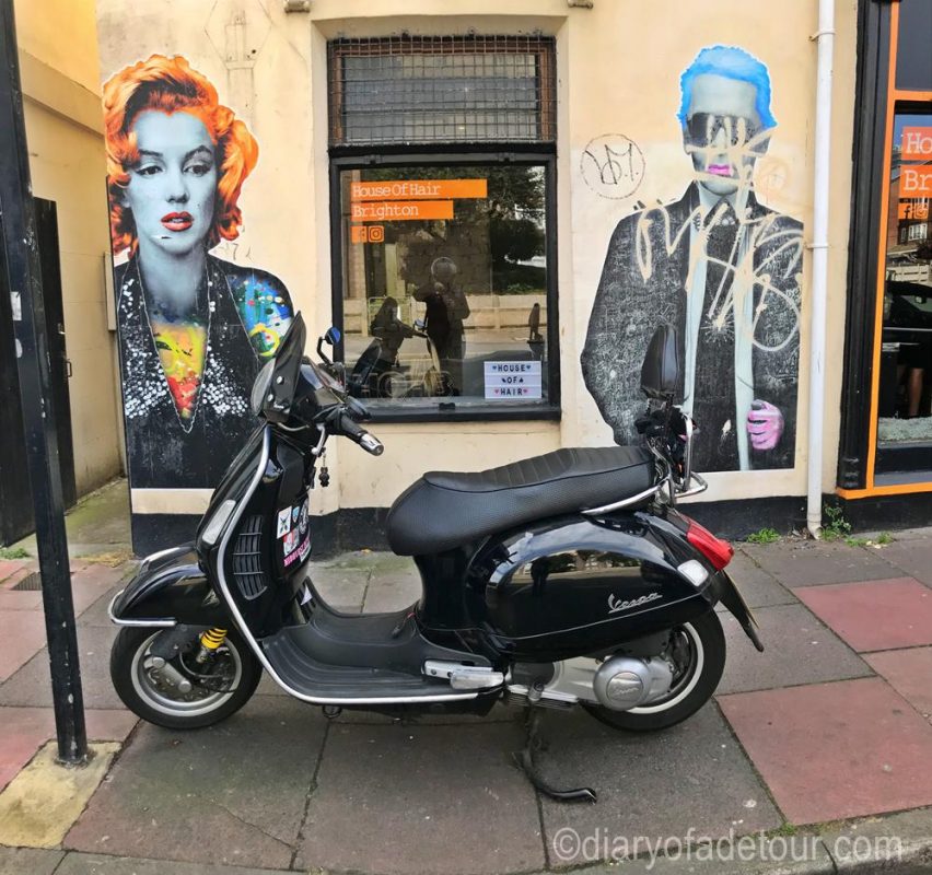 Black scooter in front of paste-ups by The Postman Artist in Kemptown, Brighton.  The paste-ups are of Marilyn Monroe with orange hair and Karl Lagarfeld with blue hair.