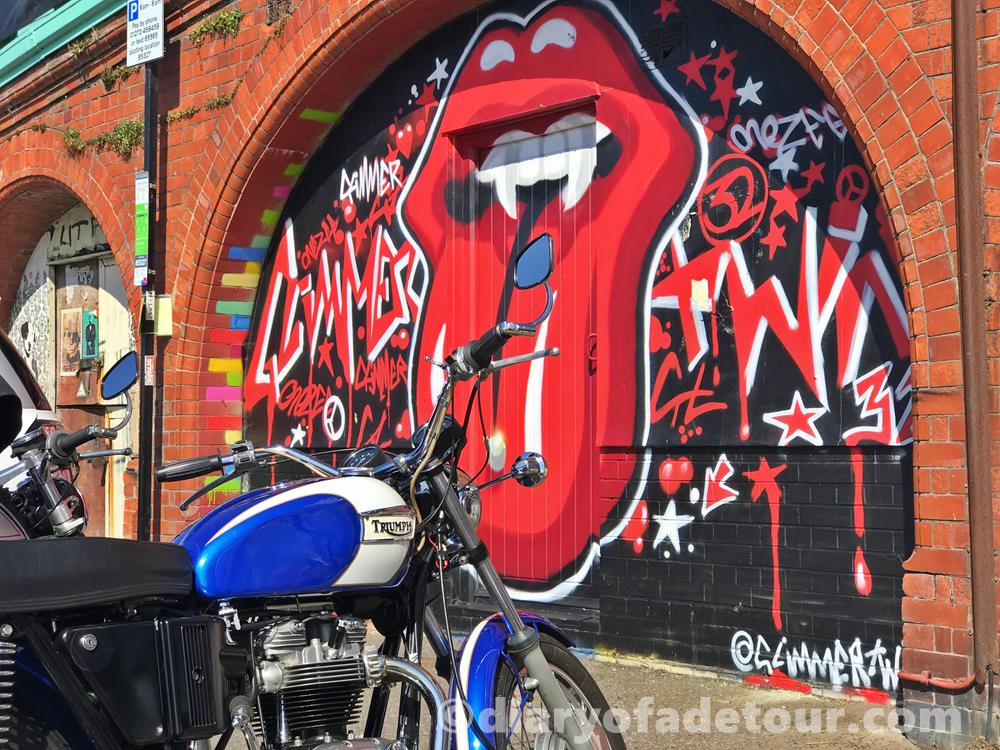 Street art mural of a tongue tagged with Glimmertwin32 in Madeira Drive, Brighton with a Triumph motorcycle in the foreground