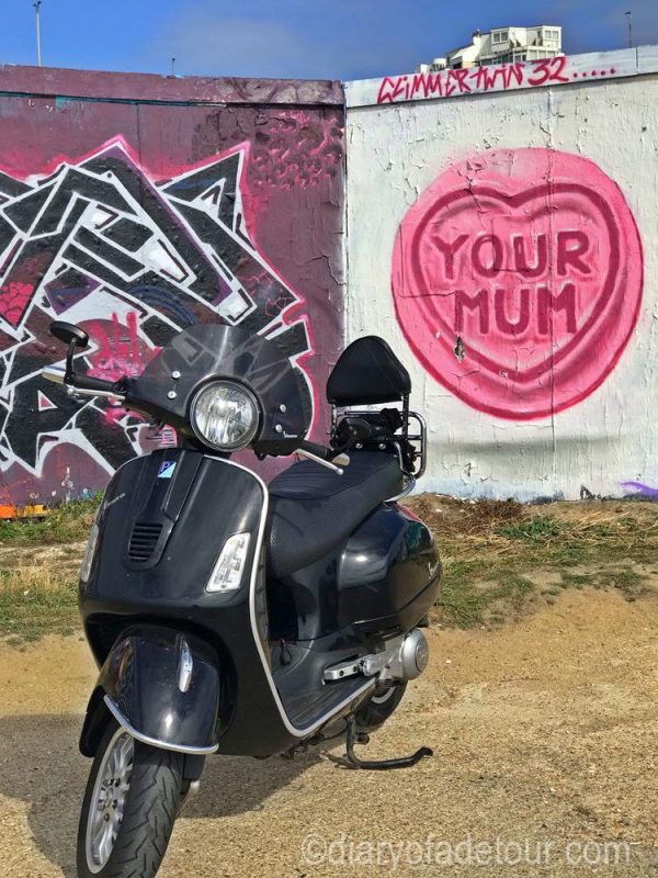 Black Vespa GTS in front of a street art mural depicting a Love Heart with Your Mum on it