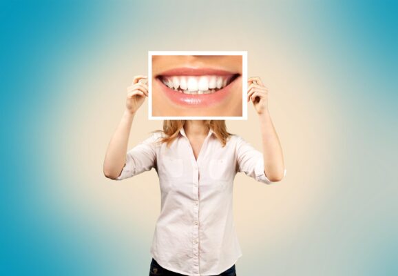 Lady in white shirt holding up a picture of a smile with sparking white teeth in front of her face