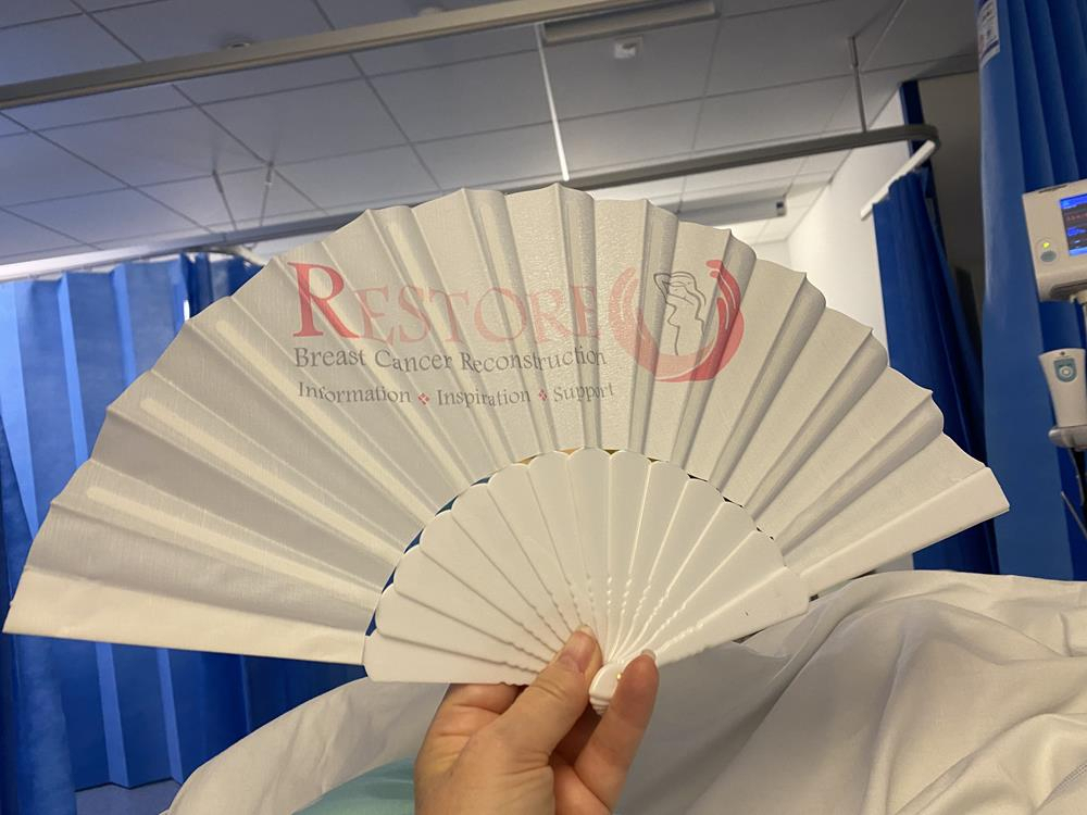Holding up a white fan in a hospital bed with the Restore Charity logo on it.